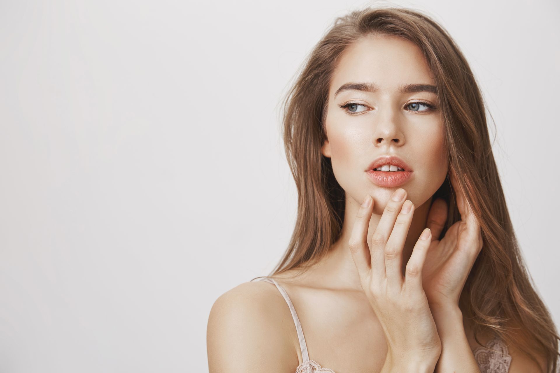 A person with long brown hair posing against a light gray background, touching their chin while looking to the side.