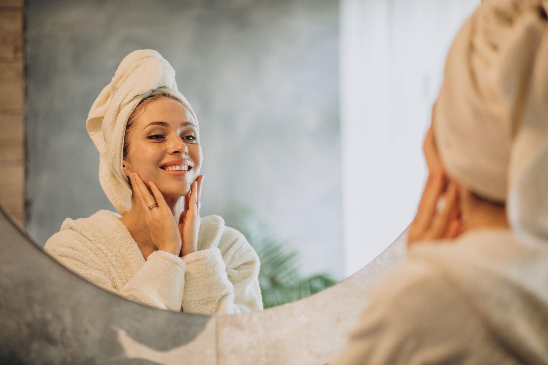 A smiling person with a white towel wrapped around their head and wearing a robe, looking into a mirror.