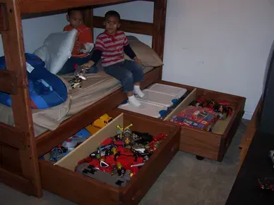 Two children are sitting on a bunk bed with drawers filled with toys.