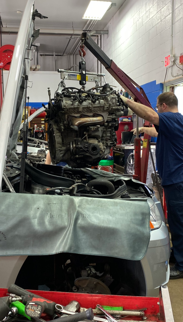 Mechanic removing a car engine with a crane in a repair shop. Silver car with open hood. | European Plus