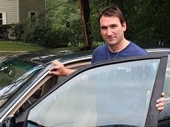 Man leans out of a car, holding the door. He wears a blue shirt and has a neutral expression. Outdoors, daytime. | European Plus