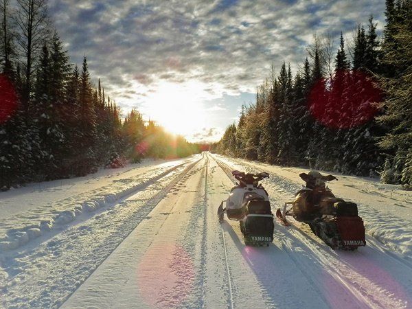 two snowmobiles on groomed trail facing a sunset