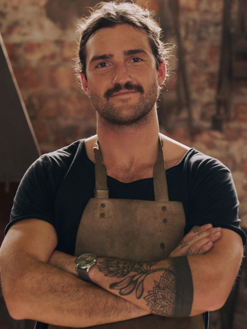 A man wearing an apron and a black shirt is standing with his arms crossed.