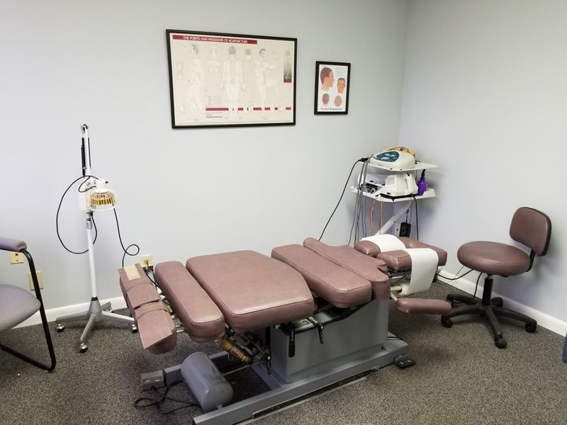 Chiropractor's treatment room with a padded table, equipment, and a stool. Gray walls and medical charts.