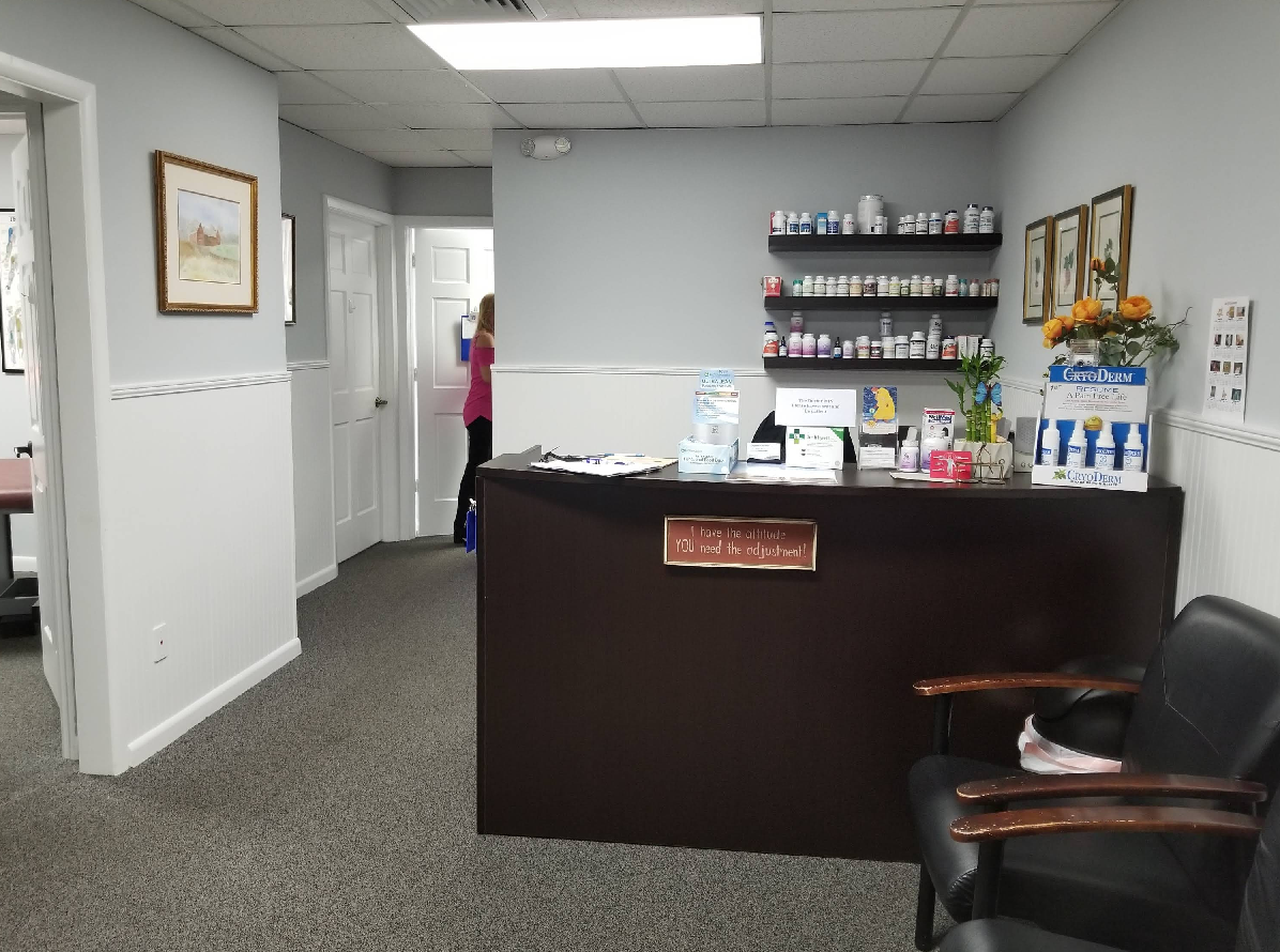Reception area of a medical office with a dark desk, shelves of medications, and a waiting chair.