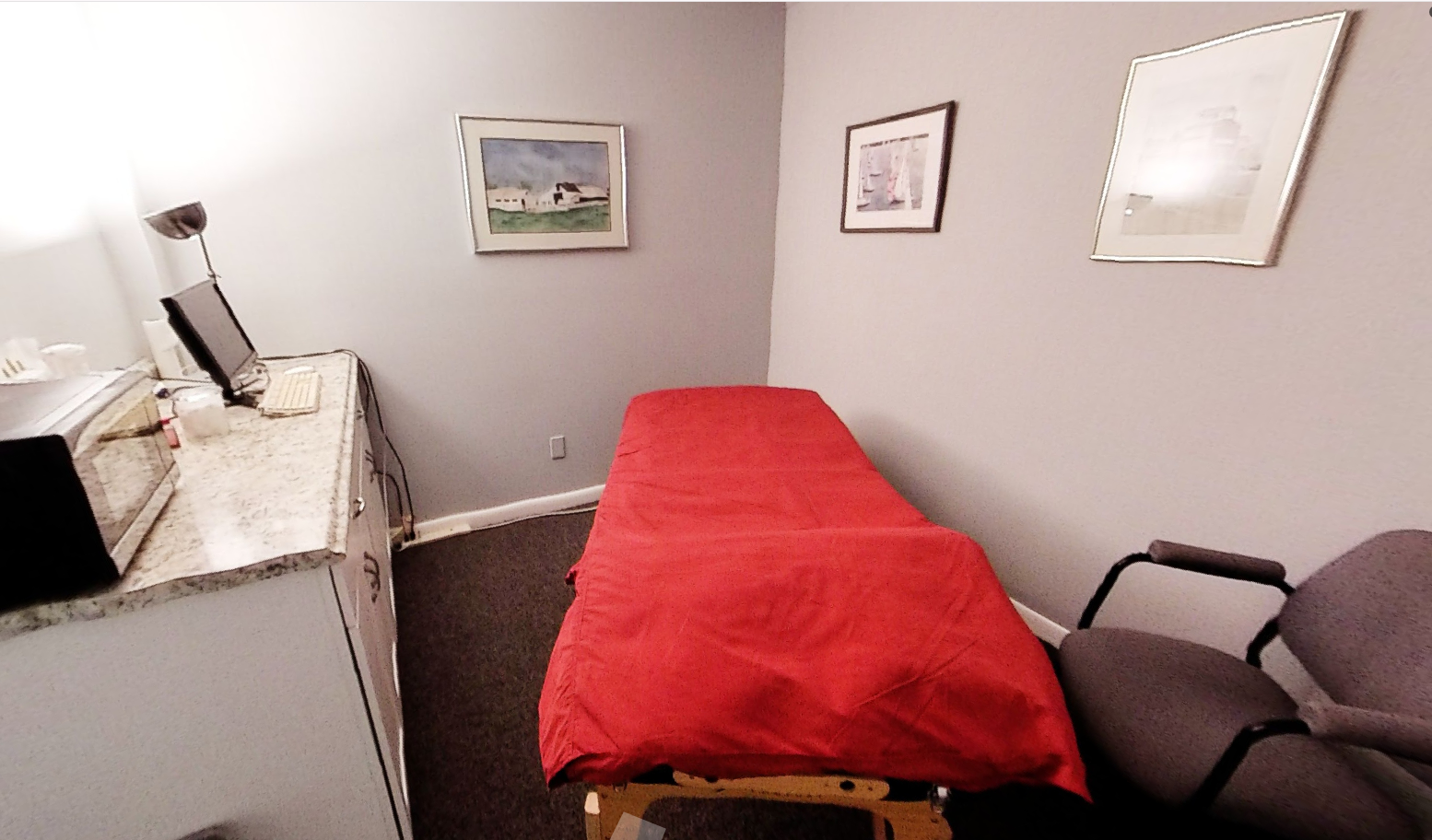 Examination room with red-covered table, gray walls, artwork, and a desk with a computer.
