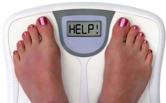 Digital scale with 0.0 reading, apple, measuring tape, and yellow dumbbells, on a wooden floor, suggesting weight loss and fitness.