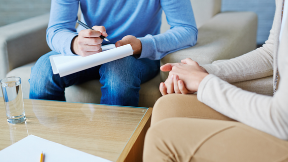 Person in blue shirt taking notes, facing someone with clasped hands, in a therapy session.