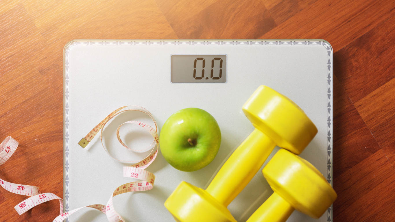 Digital scale with 0.0 reading, apple, measuring tape, and two yellow dumbbells on a wooden floor.