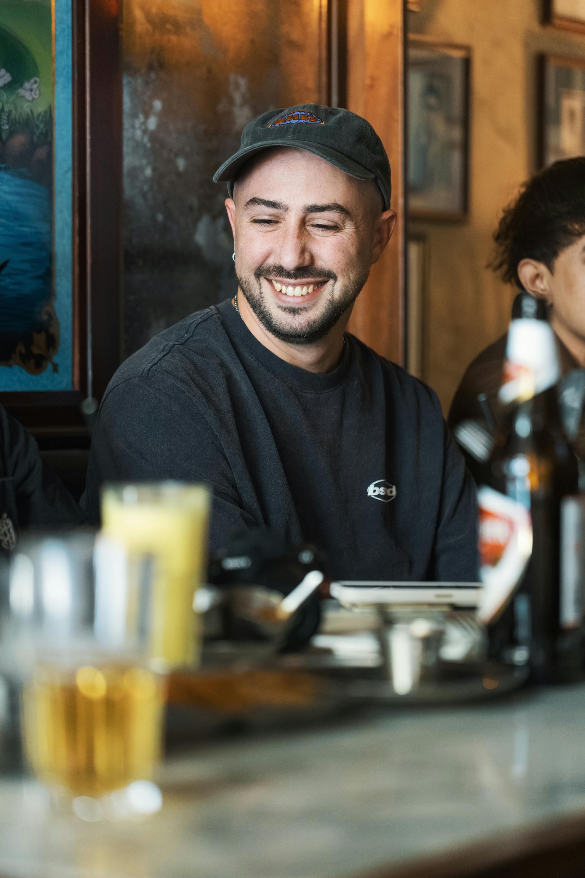 Man smiling, wearing a hat and sweatshirt, at a bar with drinks.