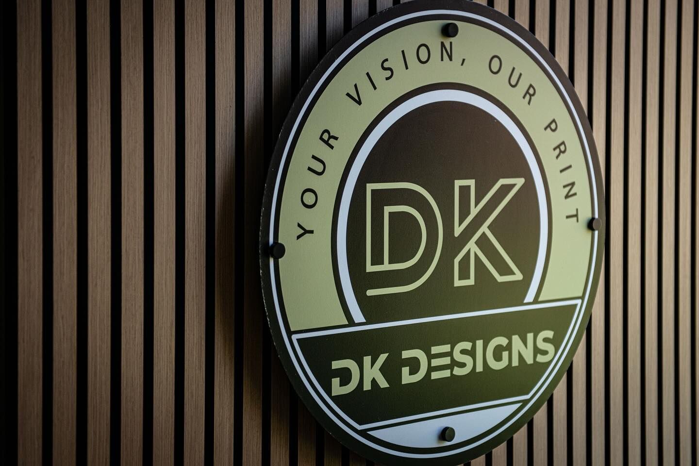 DK Designs logo on a wooden wall, gold and black circular sign. Text reads 