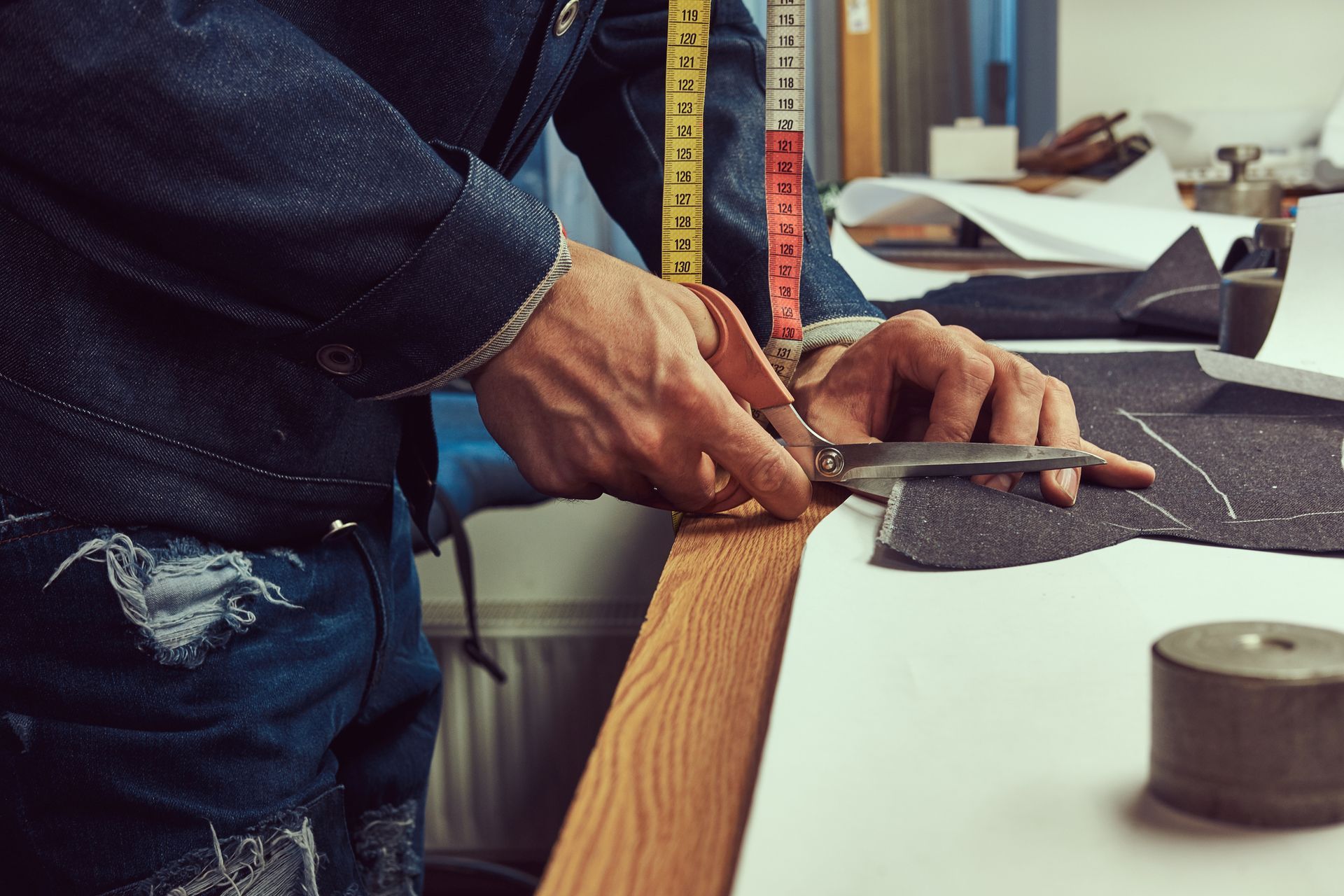 Tailor's hands using a ruler and cutter to measure and cut fabric on a white worktable.