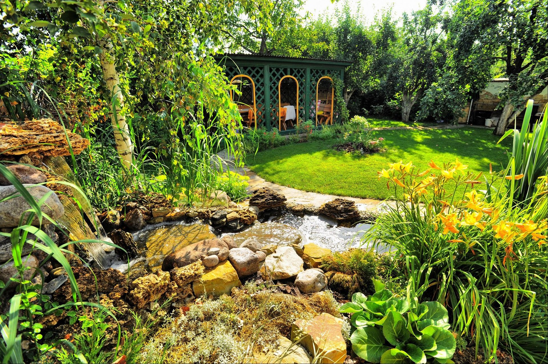 A green garden building seen behind a willow tree in a garden with a rockery and waterfall
