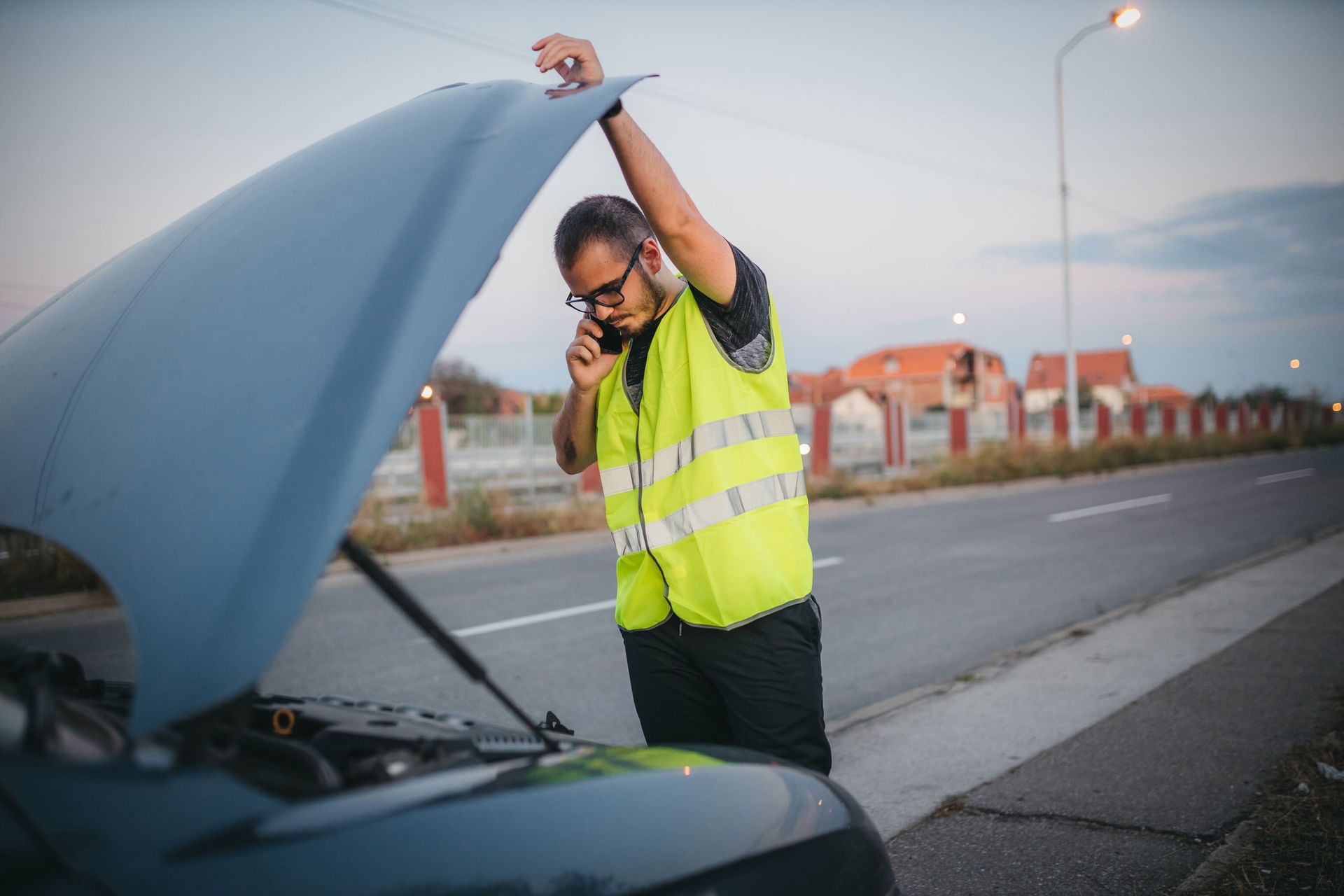 A man is standing next to a broken down car with the hood open and talking on a cell phone.