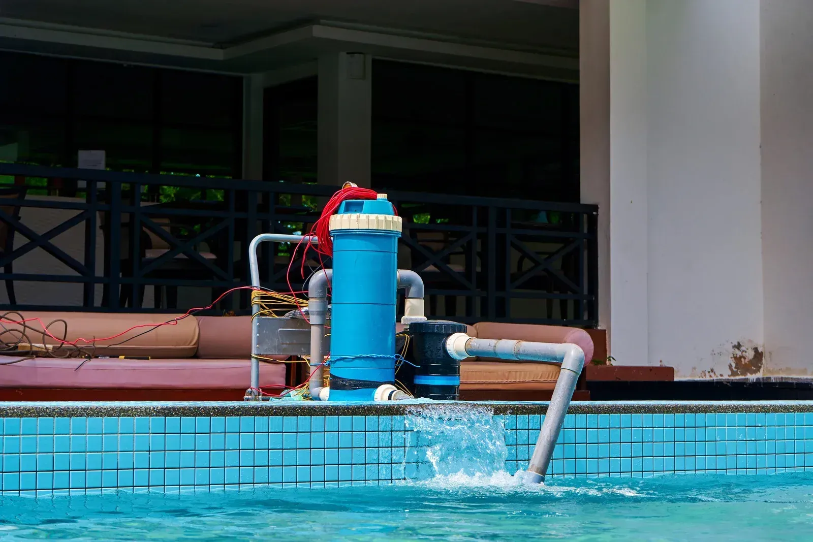 A blue pool filter unit with an attached water pipe sits poolside, actively circulating water into a tiled swimming pool.