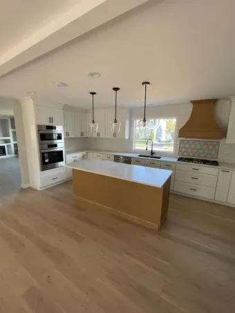 A modern kitchen with white cabinets, a central wooden island, light wood flooring, and a decorative wooden range hood.