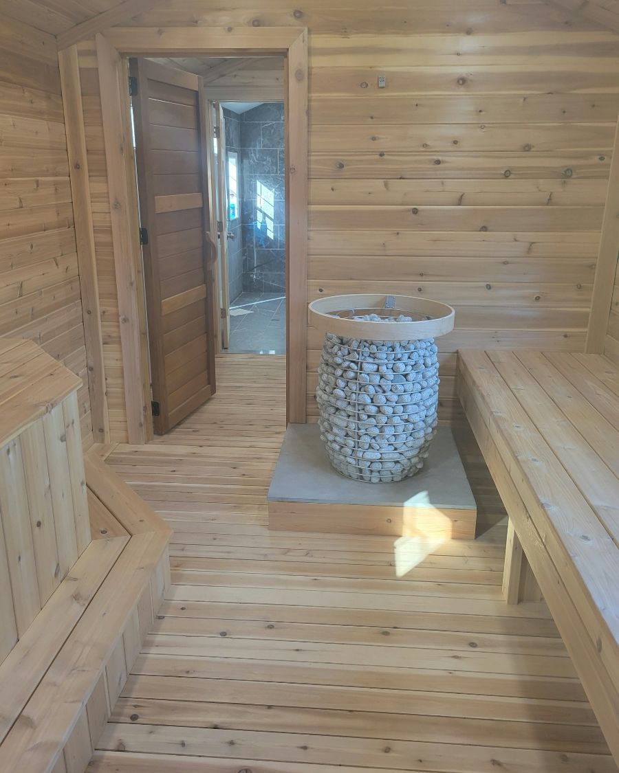 Interior of a wooden sauna with a stone-filled heater and bench seating. Open doorway to a hallway.