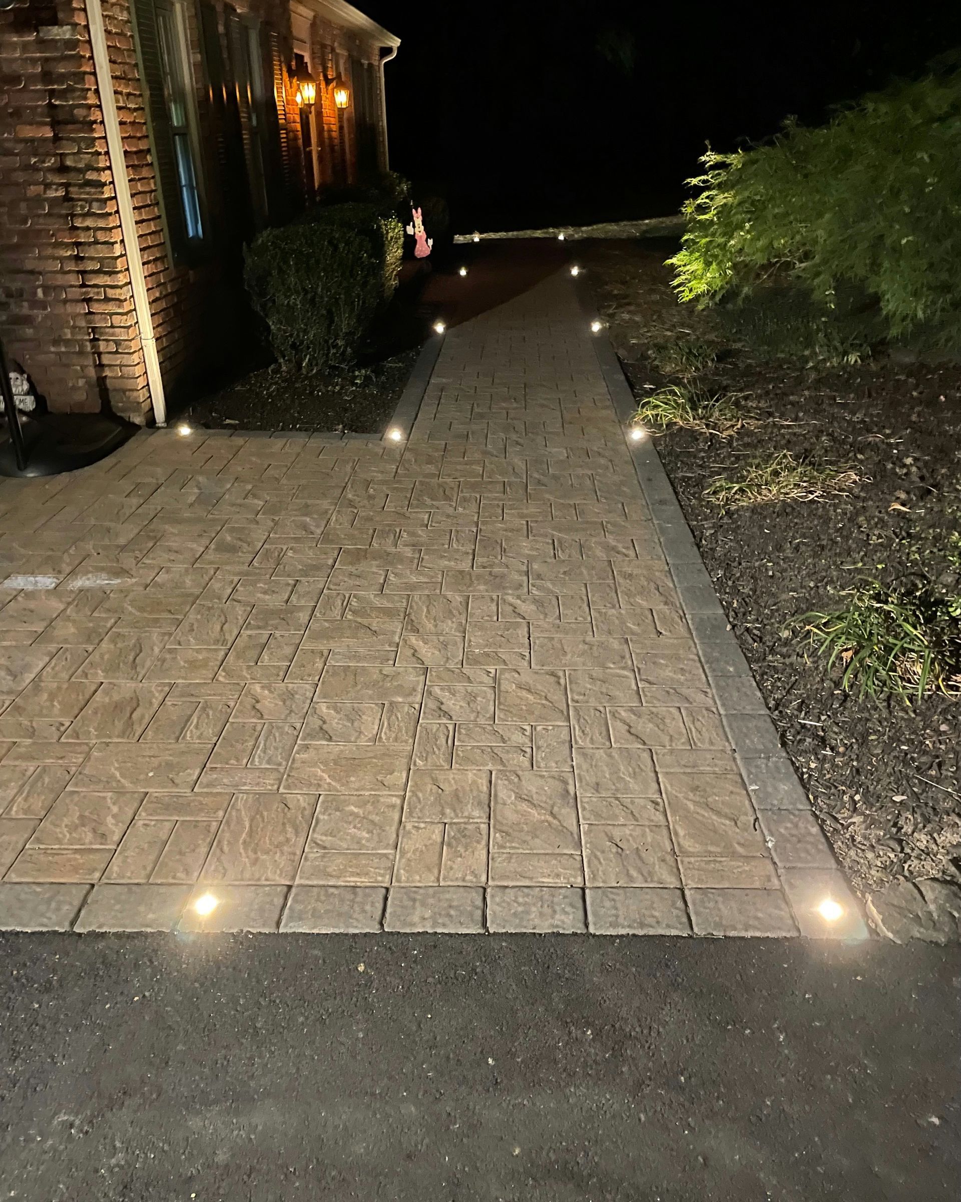 Lit walkway at night, with embedded lights along the paving stones, leading to a house entrance.