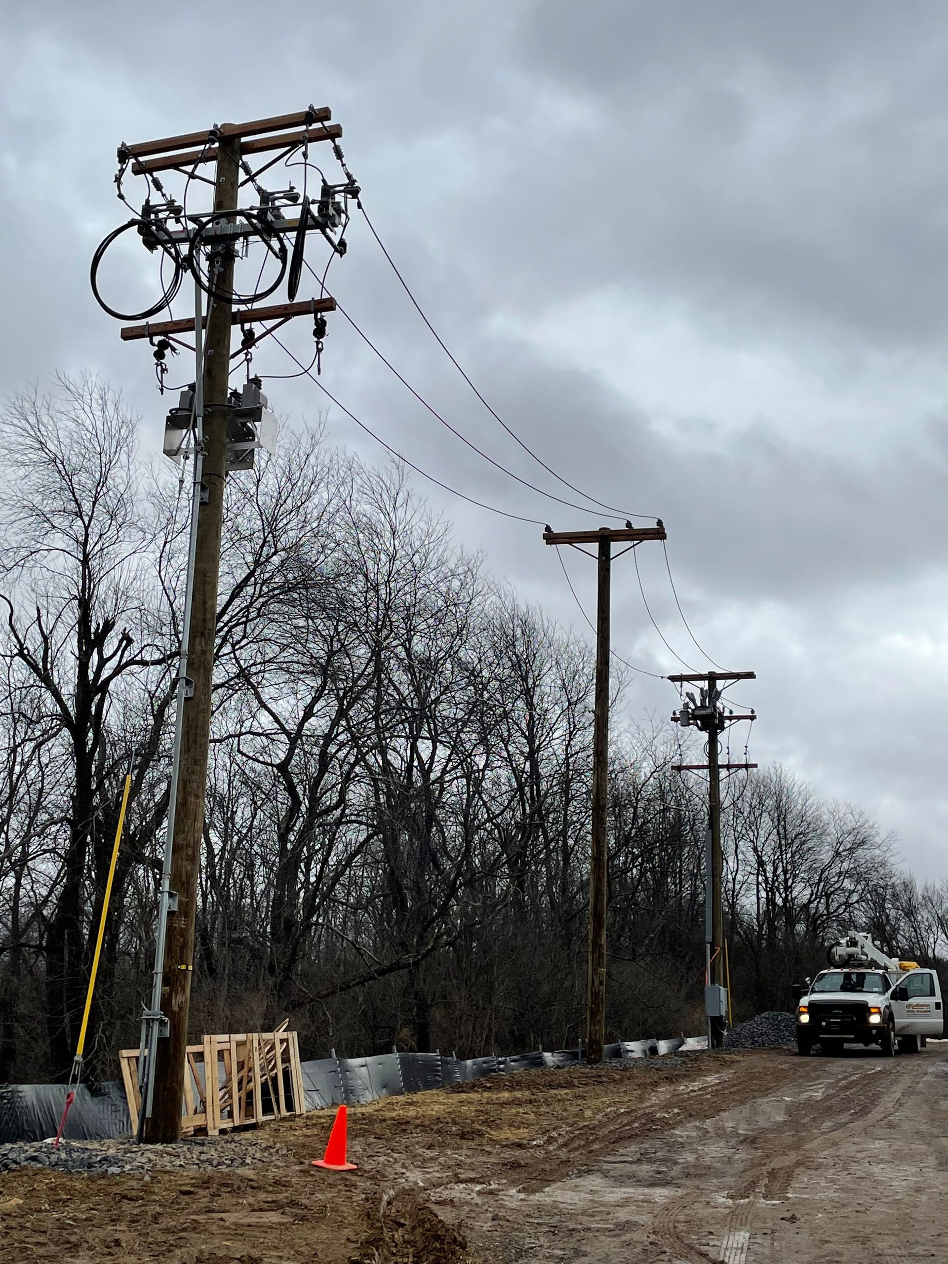 Utility poles with electrical equipment; a utility truck parked on a muddy road; overcast day.