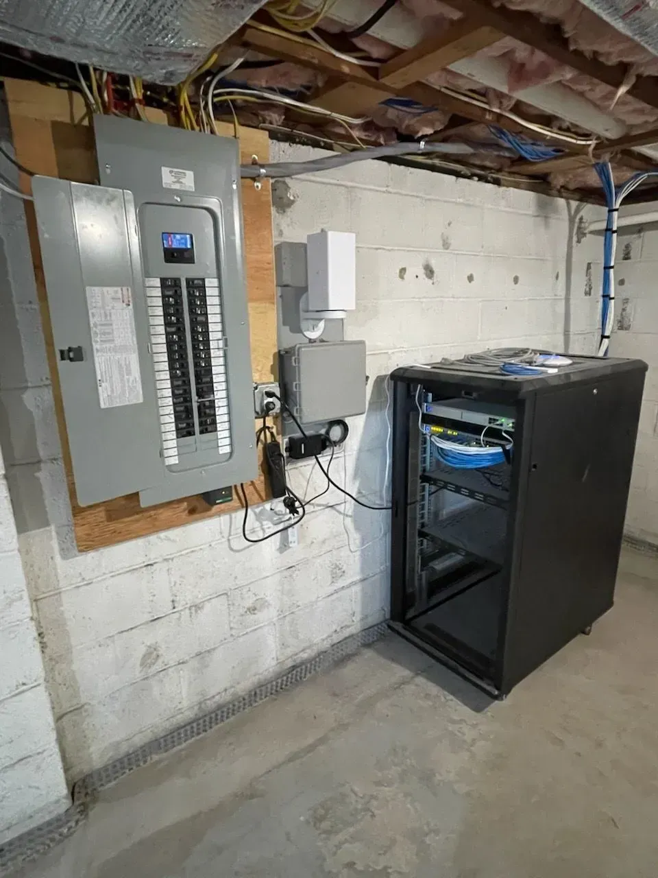 An electrical service panel and a black network server rack mounted on a basement concrete wall with exposed ceiling joists.