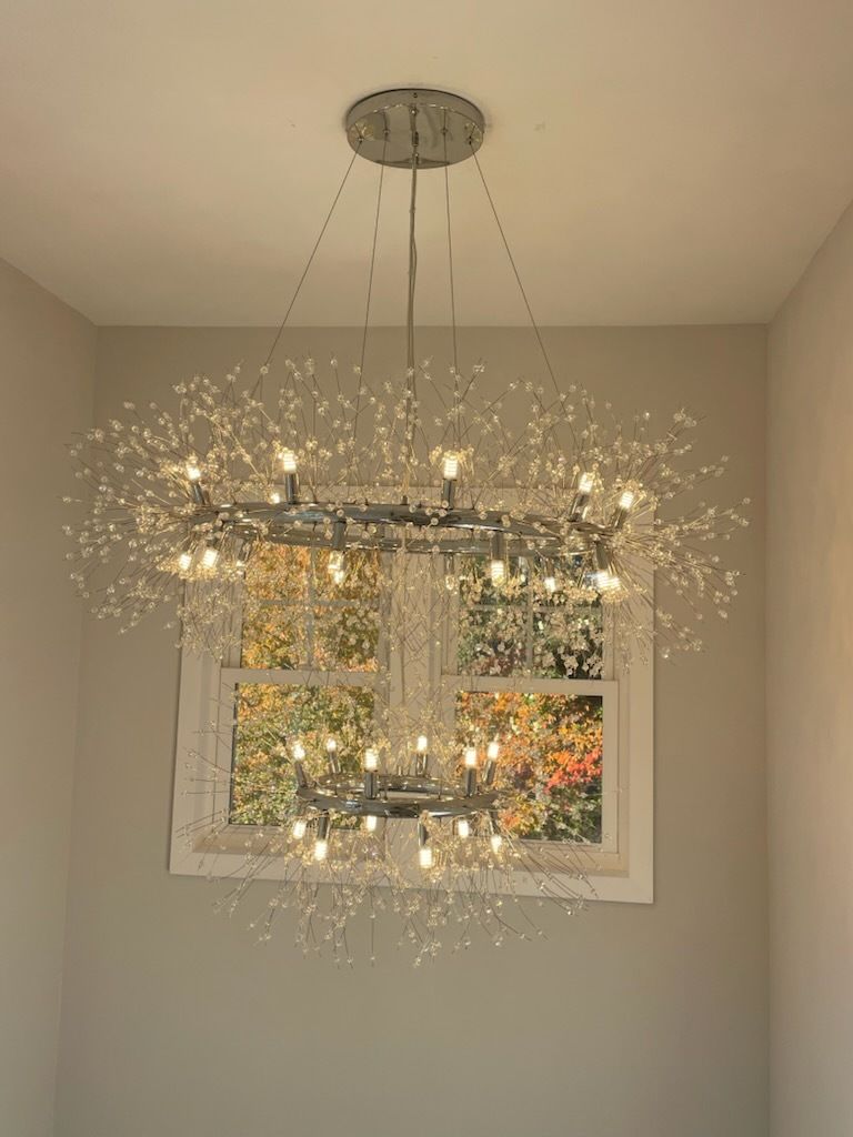 Modern chandelier with multiple lights, hanging in front of a window with autumn foliage visible.