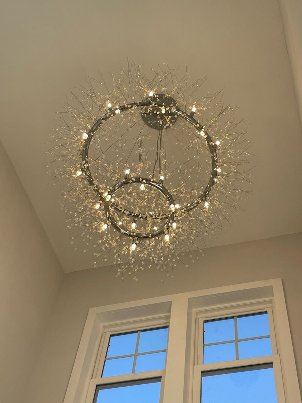 A modern chandelier with multiple circular light fixtures. It's mounted on a white ceiling above two windows.