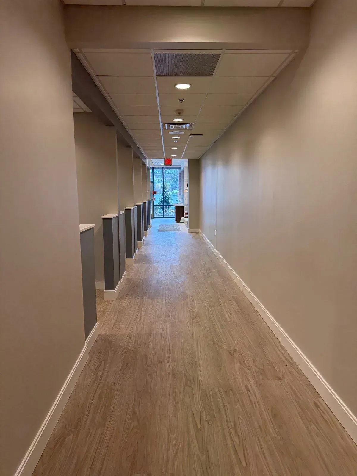 A long, brightly lit hallway with light wood-style flooring, gray partition walls on the left, and solid walls on the right.