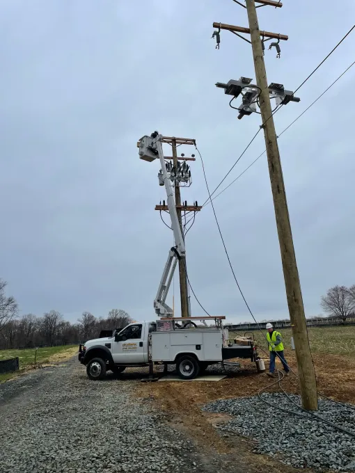 A utility worker in a high-visibility vest stands near a bucket truck working on utility poles in a grassy field.