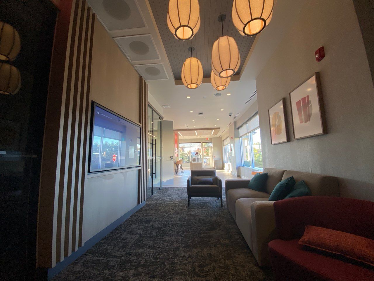 Lounge area with gray carpet, brown couch, art, TV, pendant lights. View of hallway and entrance.