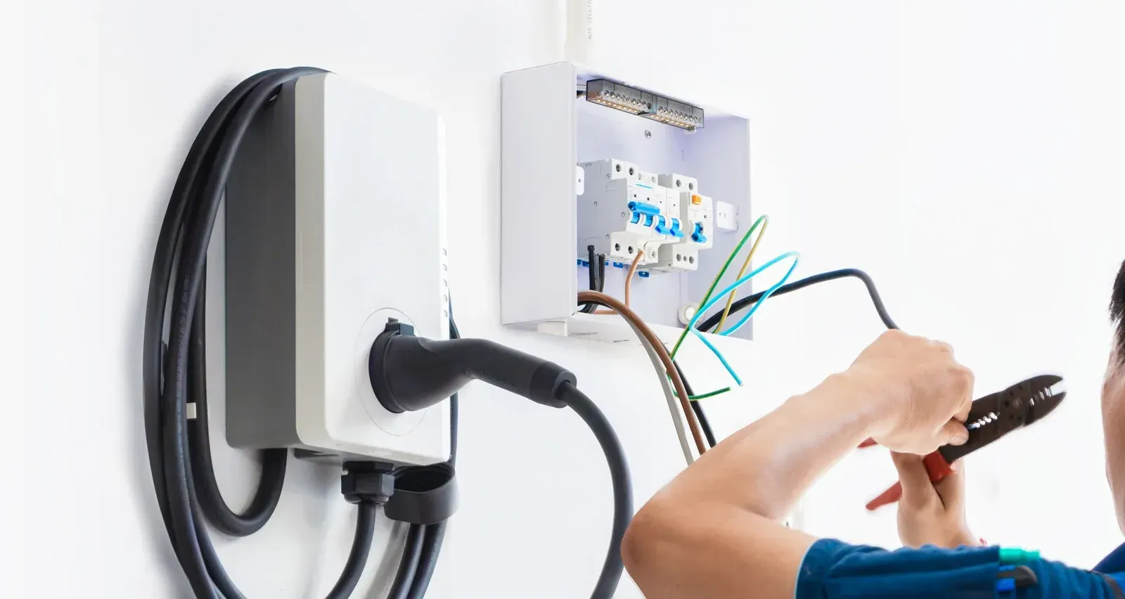 An electrician uses wire cutters to install an electric vehicle wall charger in a white electrical box.