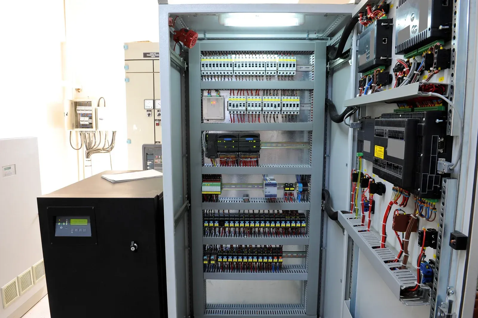 Open electrical control panel with circuit breakers, wires, and a nearby black UPS unit in a server room.