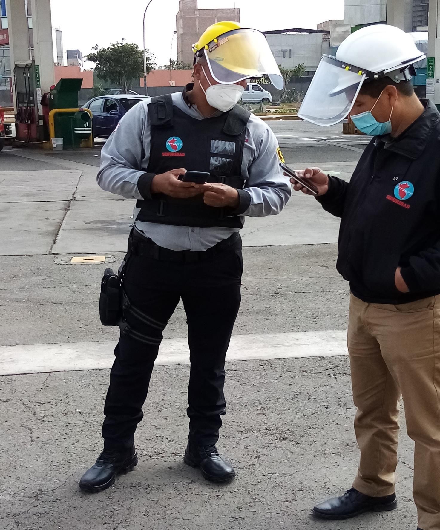 Dos personas con mascarillas y protectores faciales en una gasolinera, mirando sus teléfonos. Una lleva chaleco.