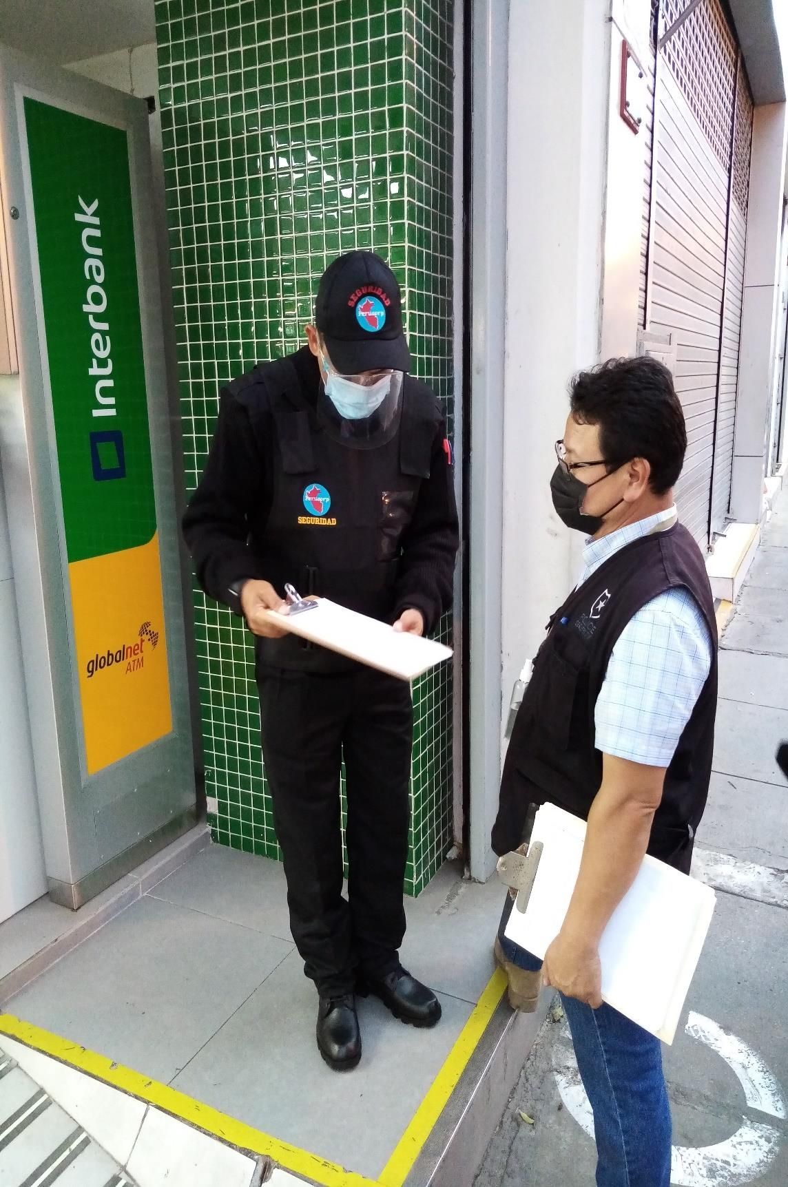 Dos personas con mascarillas se encuentran cerca de un cajero automático de Interbank. Una de ellas sostiene un portapapeles.