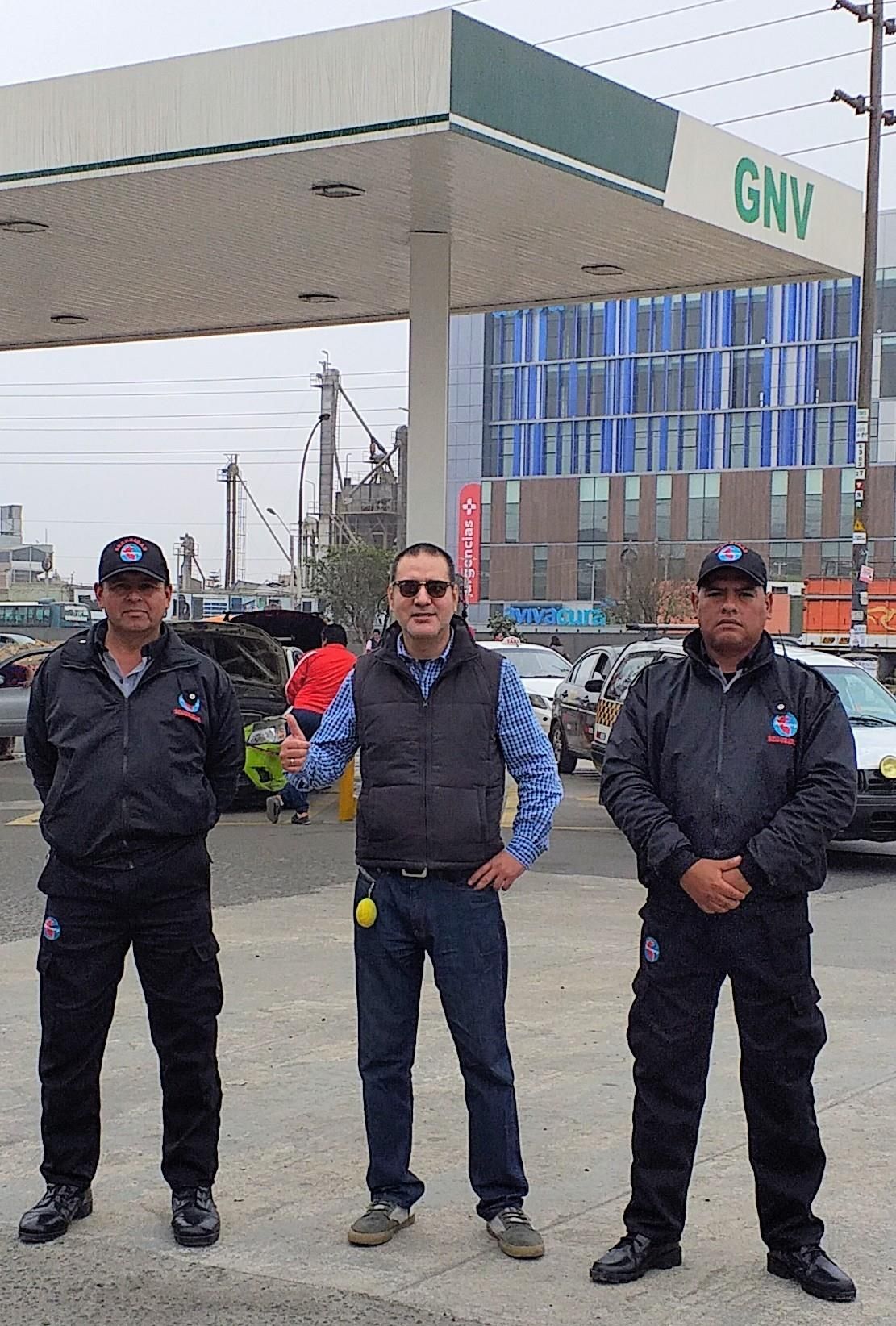 Tres hombres uniformados se encuentran frente a una gasolinera bajo un toldo; uno de ellos levanta el pulgar.