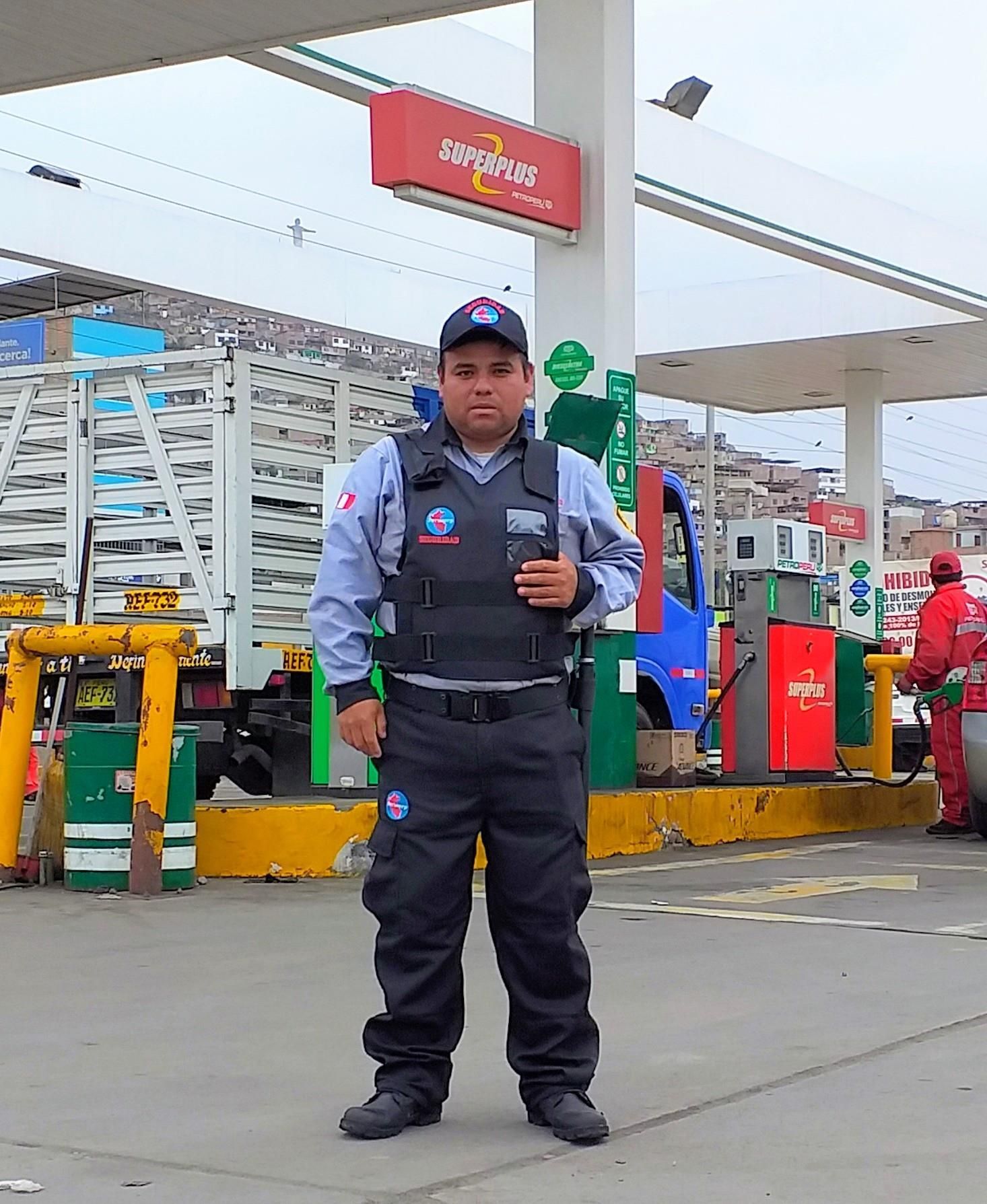 Un guardia de seguridad uniformado se encuentra en una gasolinera, sosteniendo una radio, con un camión y bombas al fondo.