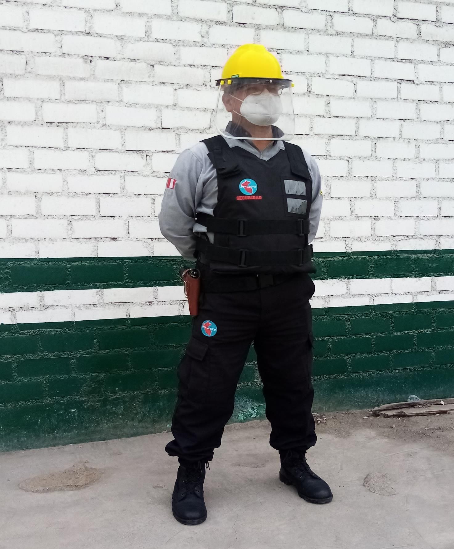 Persona con casco amarillo, máscara y chaleco antibalas, de pie frente a una pared de ladrillos blancos con detalles verdes.