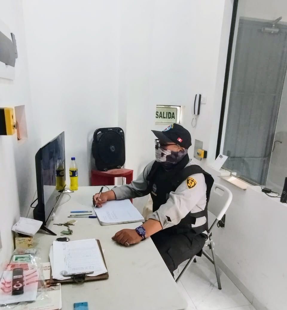 Persona uniformada sentada en un escritorio, escribiendo. Lleva mascarilla y se encuentra en una pequeña habitación blanca.