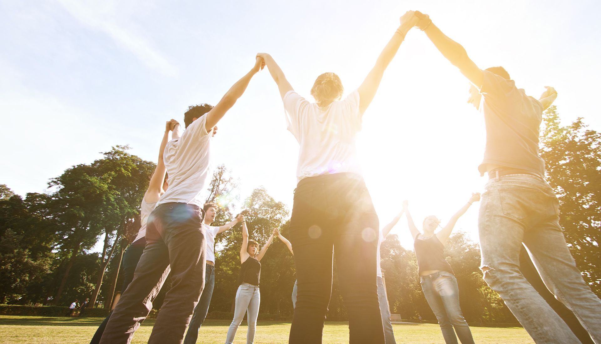 Group of people holding hands in a circle outdoors with arms raised in a sunny park.