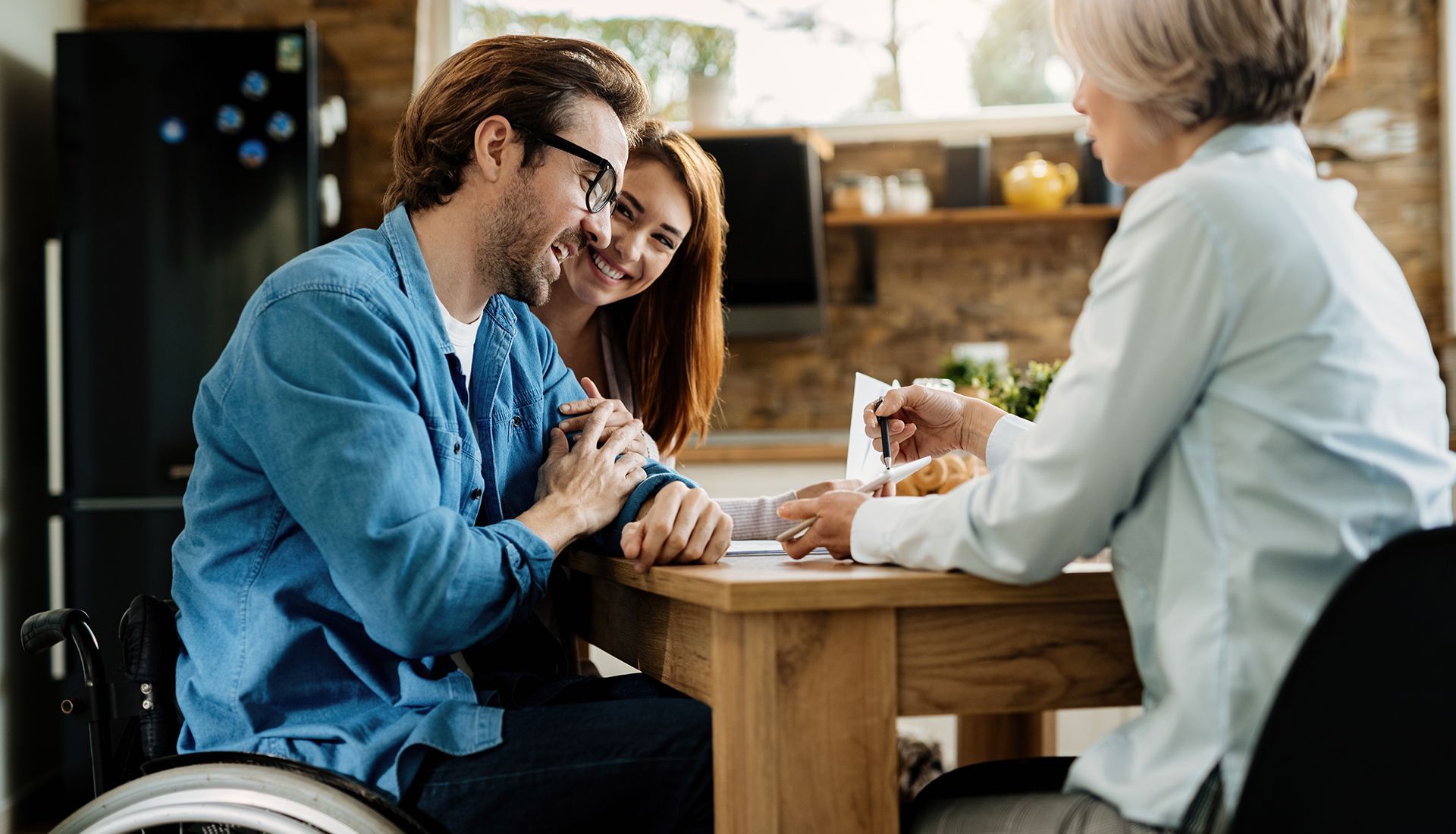 Couple seated at a table with a woman, possibly a consultant, in a kitchen setting. The man is in a wheelchair.