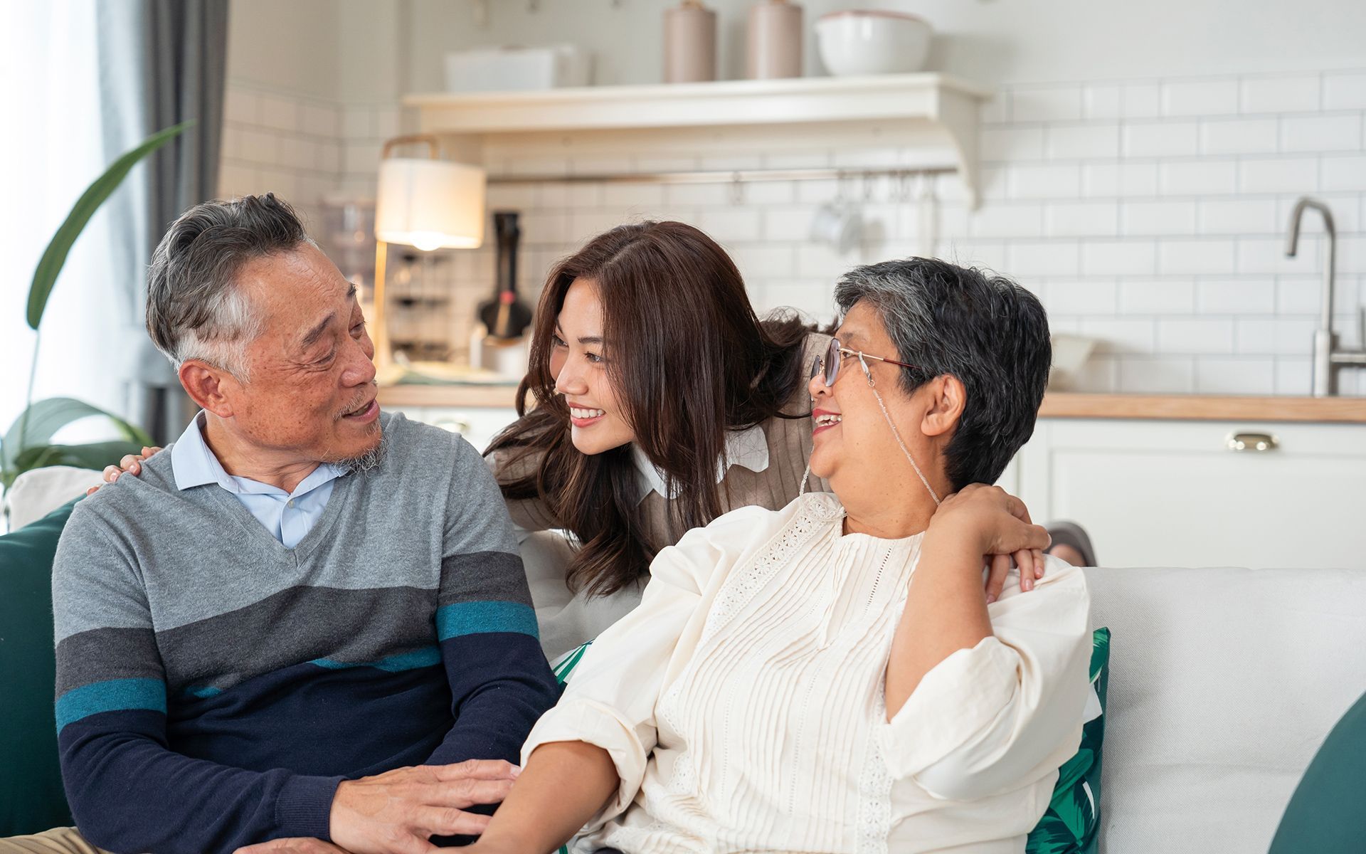 A young woman embraces an elderly couple on a couch in a well-lit living room.