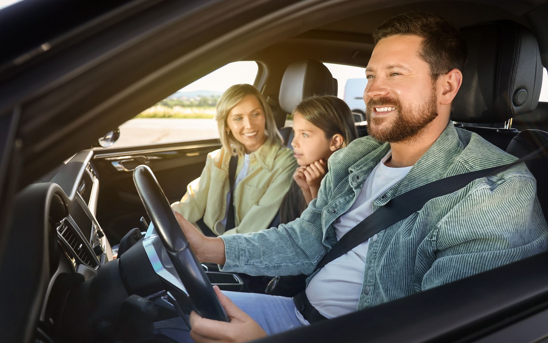 Family of three smiling in a car: Father driving, mother and child in the back, sunny outdoors.