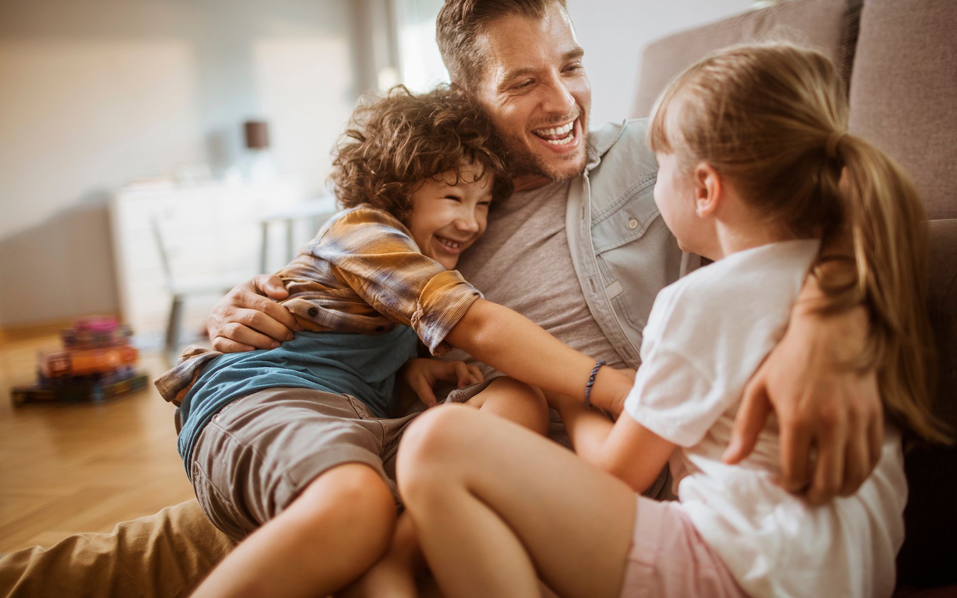 Man hugging two laughing children on a couch in a living room.