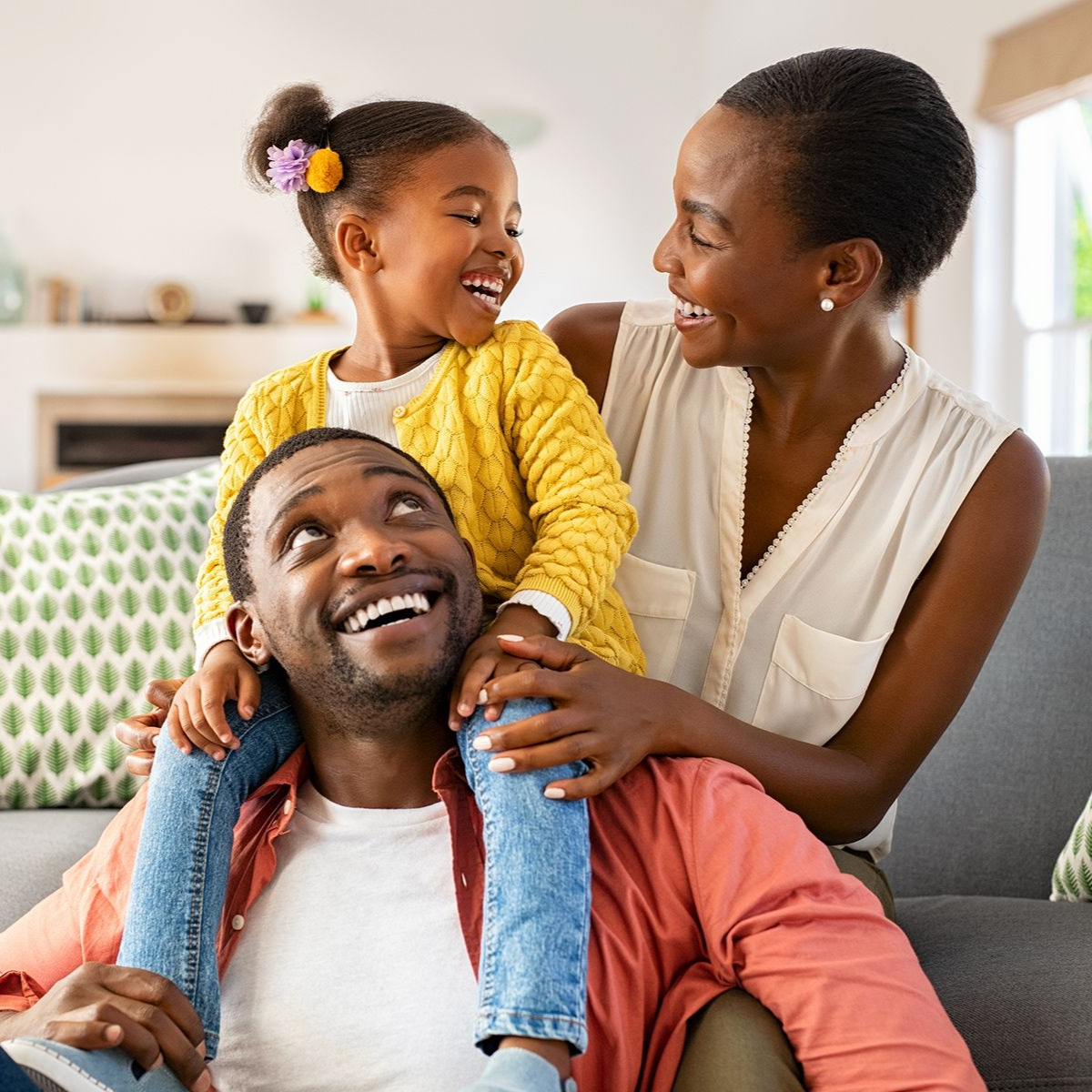 Family of three, smiling: daughter on father's shoulders, mother beside them on a couch indoors.