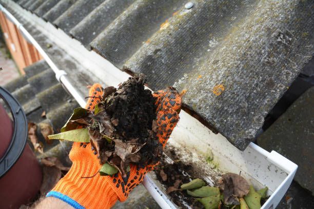 Person wearing orange gloves cleaning a gutter filled with leaves and debris on a rooftop.