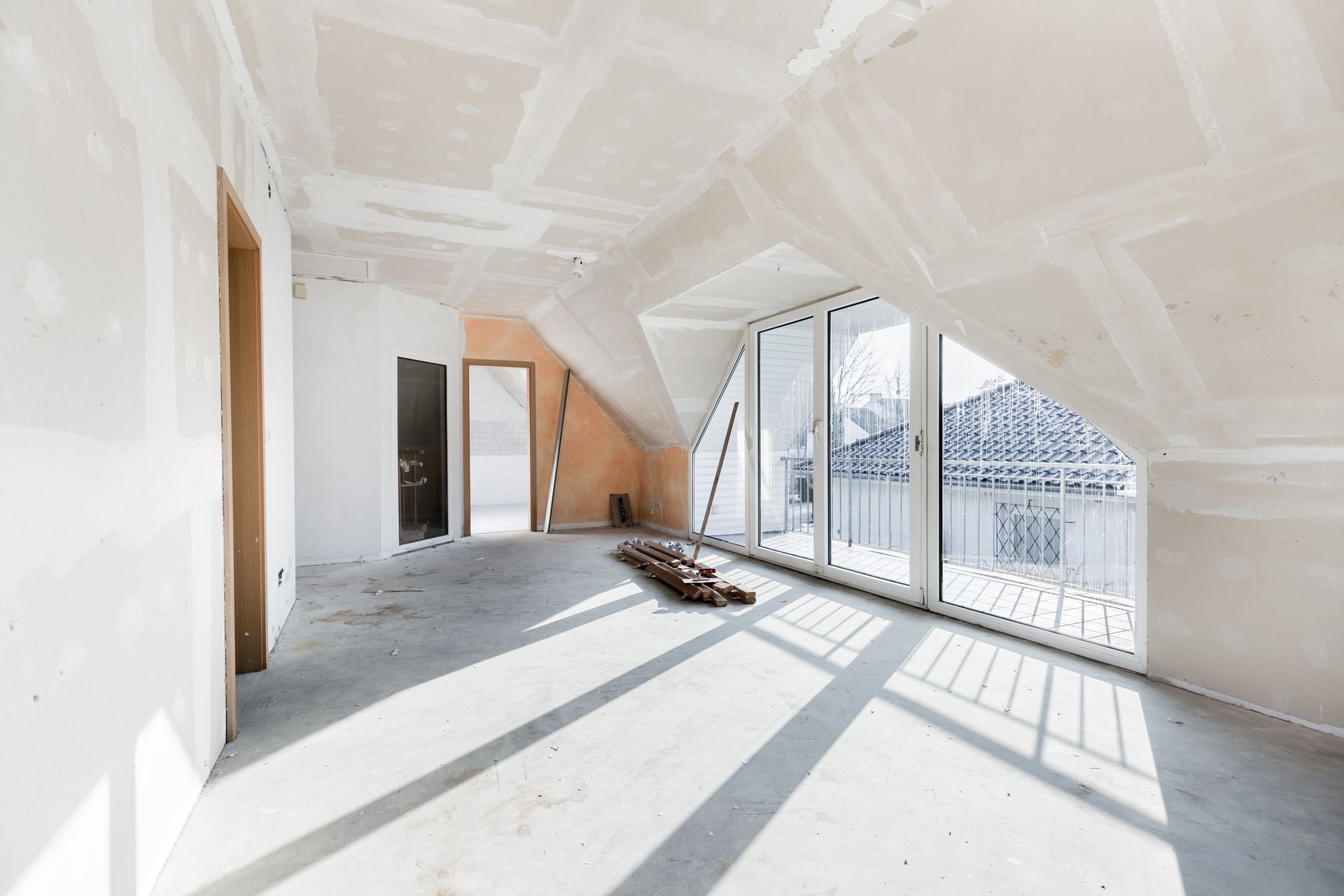 Interior of a room under construction, with drywall and large windows. Sunlight casts shadows on the floor.