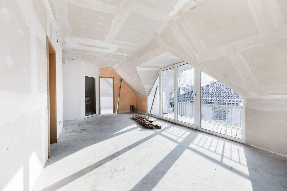 Interior of a room under construction, with drywall and large windows. Sunlight casts shadows on the floor.