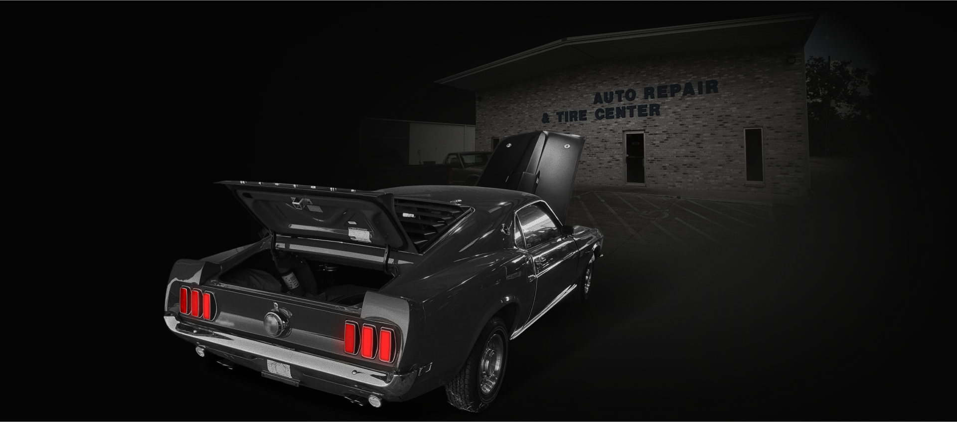 A black vintage car with an open trunk in front of an auto repair shop at night. | Deville Automotive