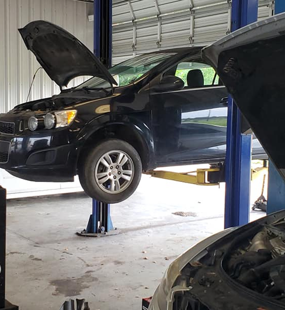 Black car raised on a lift in a garage, hood open, being worked on. | Deville Automotive