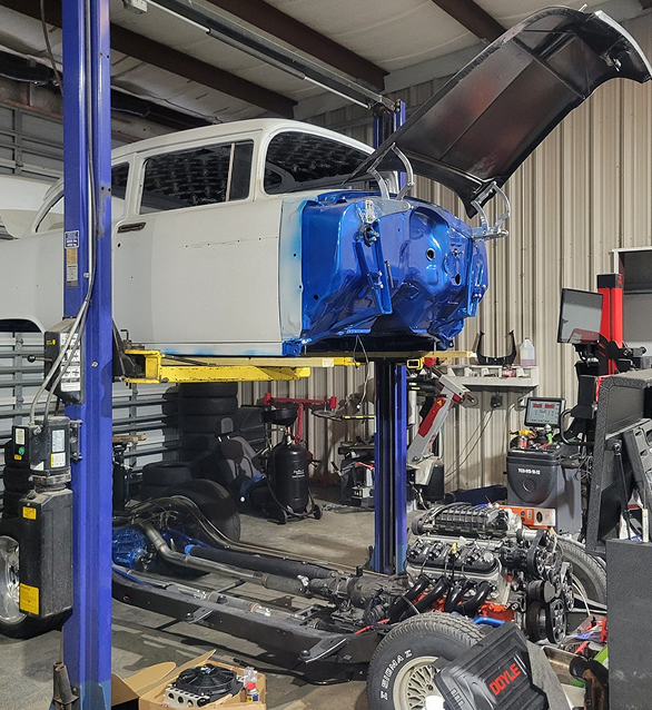 Car body on a lift in a shop, undergoing restoration. Blue and white paint, open hood, chassis visible. | Deville Automotive