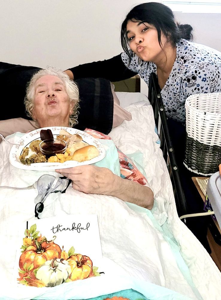 A woman is kissing an elderly woman in a hospital bed while holding a plate of food.